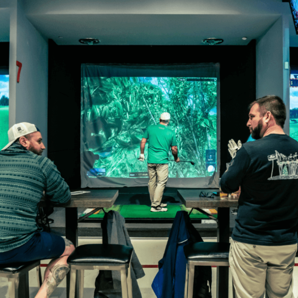 Two Males Sitting at Bar Tables and Chairs Behind Golfer on TwoVision Simulator in Bay 7