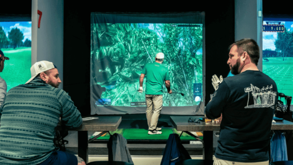 Two Males Sitting at Bar Tables and Chairs Behind Golfer on TwoVision Simulator in Bay 7