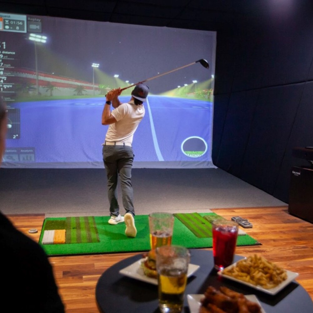 Golfer Swinging on Moving Swing Plate with Beer and Food on Bar Table Behind Simulator