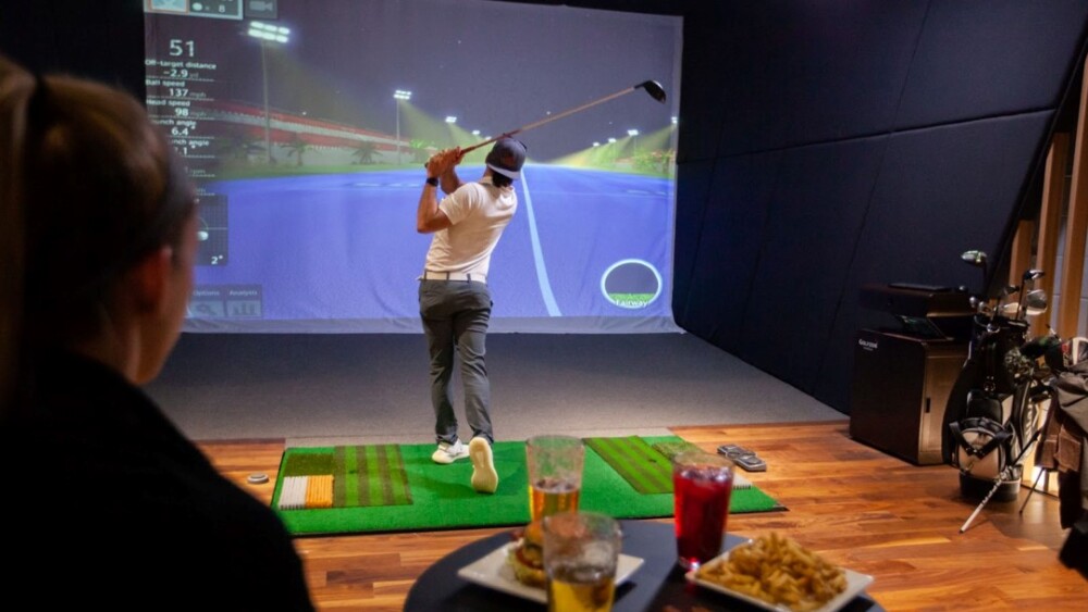 Golfer Swinging on Moving Swing Plate with Beer and Food on Bar Table Behind Simulator